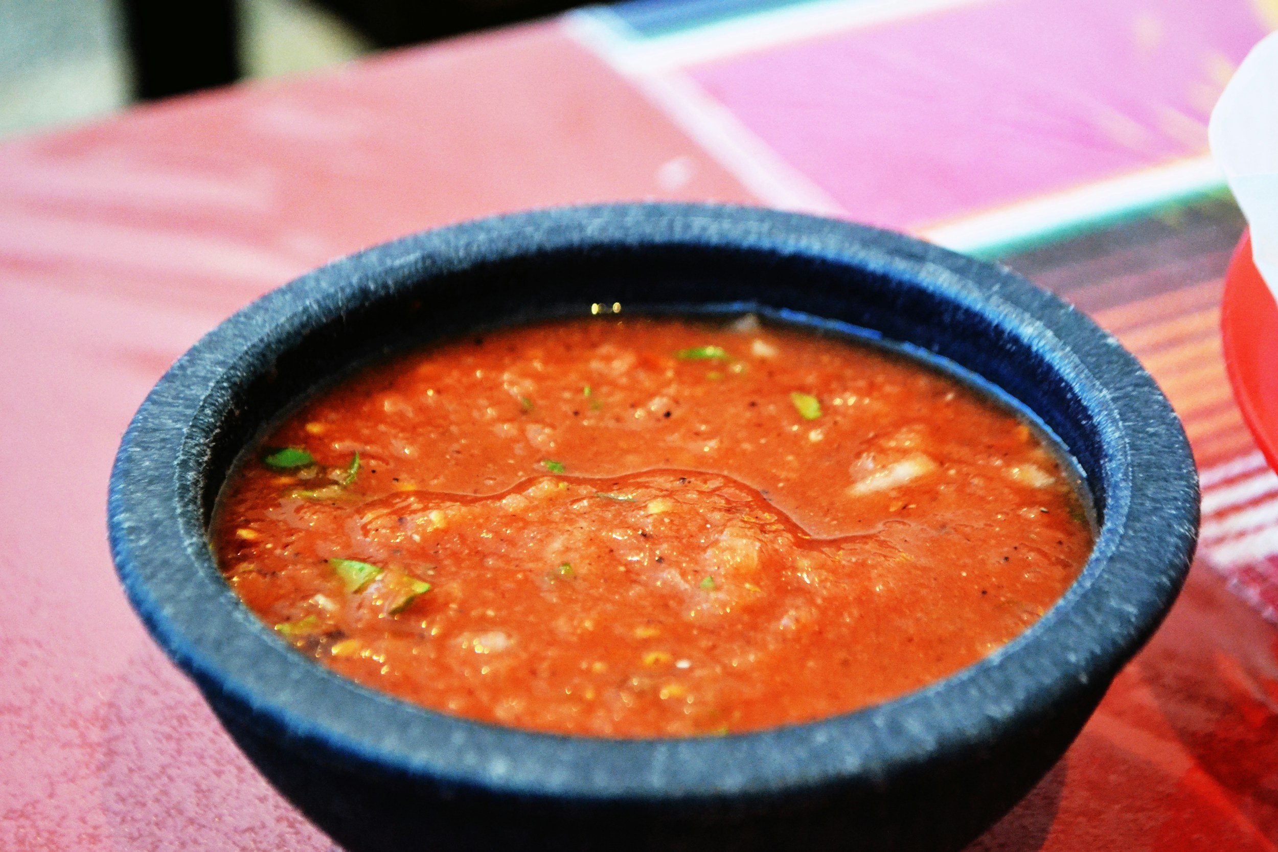 Bowl of fresh salsa served in a traditional stone dish, showcasing a rich red color with visible herbs and spices, placed on a colorful table setting.