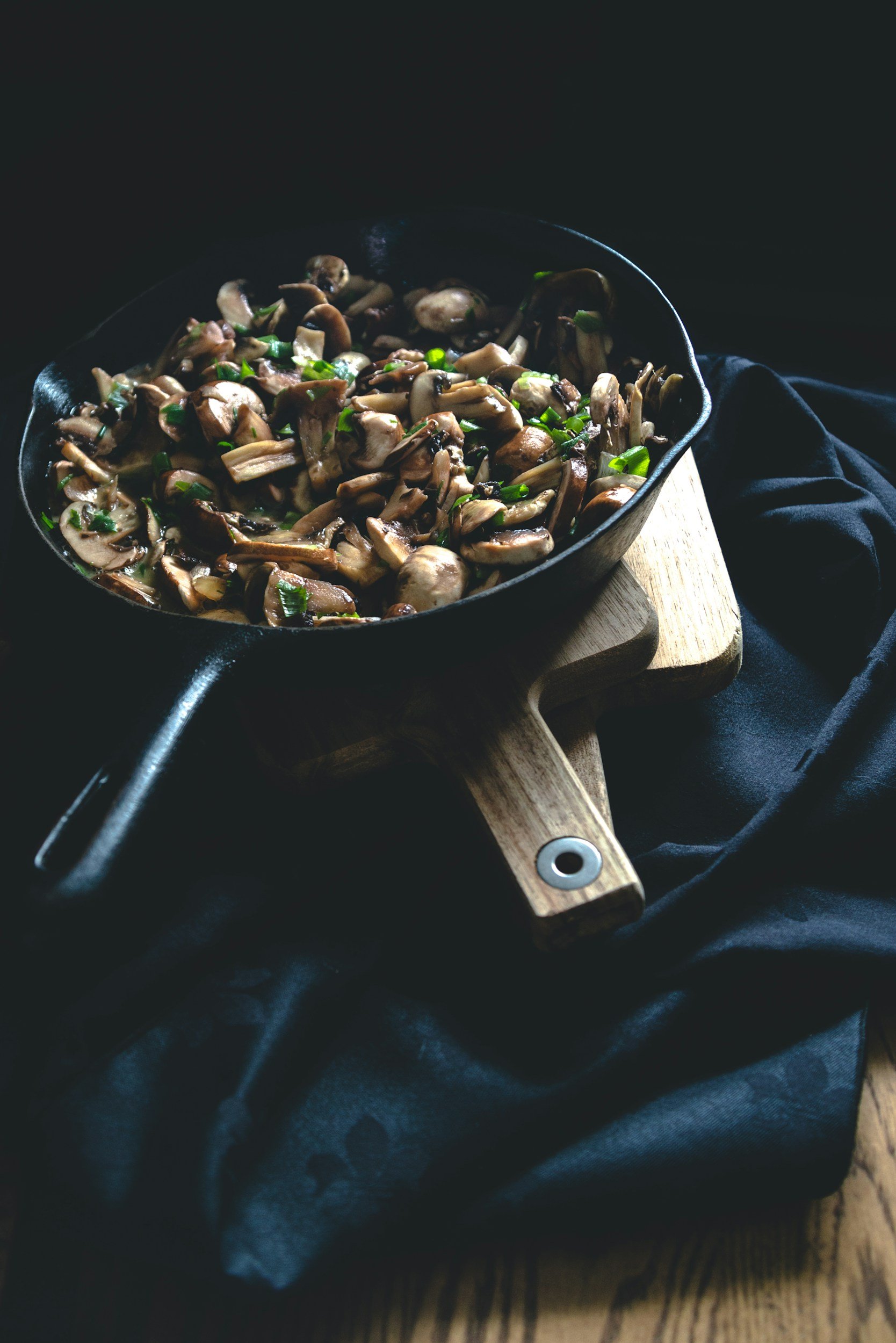 Sautéed mushrooms in a cast iron skillet garnished with green herbs, placed on a wooden cutting board over a dark fabric background.
