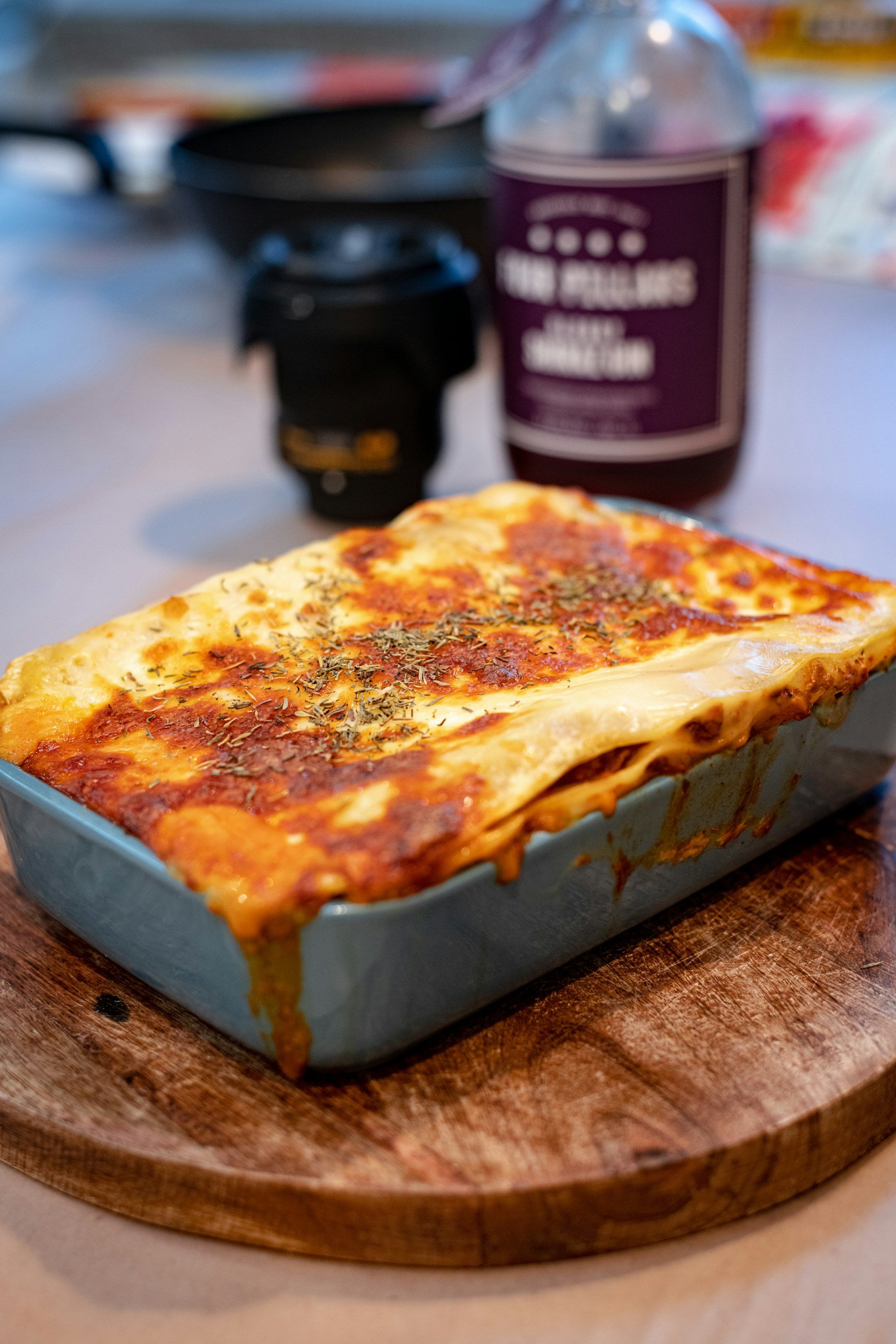 Baked lasagna in a blue ceramic dish, topped with melted cheese and herbs, placed on a wooden cutting board, with a blurred background featuring a black coffee cup and a bottle of beverage.